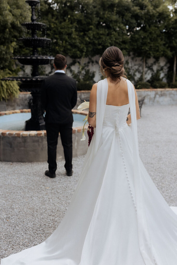A bride and groom in the courtyard of the Supposey Warehouse and Gardens wedding venue, ready for their first look. The bride is standing behind the groom, who is facing away from her.