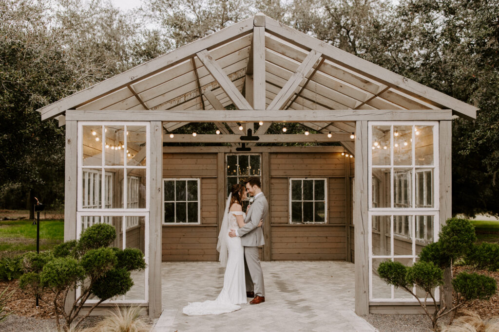 A couple standing in front of the greenhouse at the Mill Pond Estate wedding venue, facing each other with their arms around each other's waists.