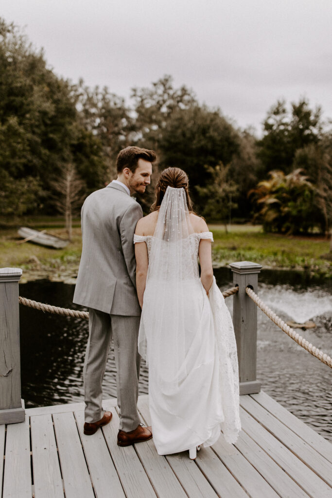 A couple standing on the dock of the lily pond at Mill Pond Estate wedding venue. They are looking away towards the water, side by side. 