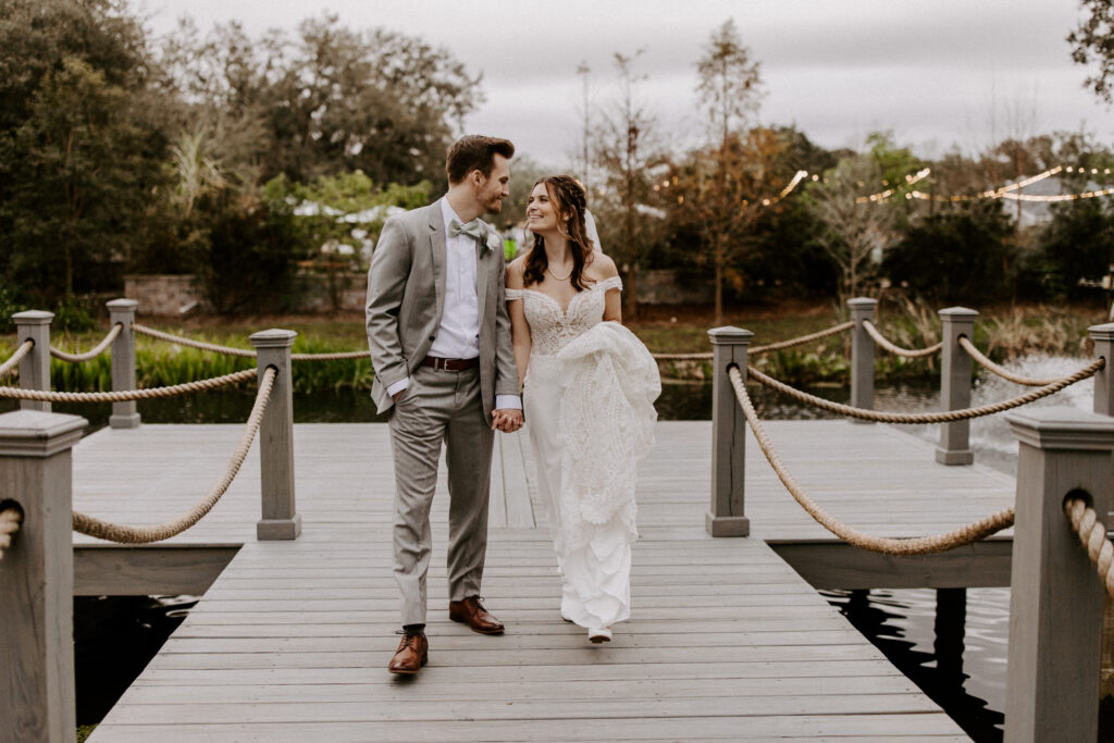 A couple walking side by side, on the dock at the Mill Pond Estate wedding venue. They are holding hands, looking at each other as they walk.
