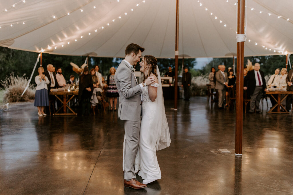 A couple dancing under the sailcloth tent at the Mill Pond Estate wedding venue. They are facing each other, laughing, and holding hands. Their guests are behind them at the tables, watching. 